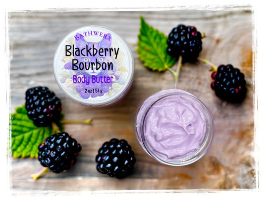 BathWerx Blackberry Bourbon Body Butter - 2 oz jar with lid and open jar showing lavender-purple whipped butter, surrounded by fresh blackberries and green leaves on rustic wood surface