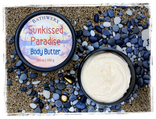 BathWerx Sunkissed Paradise Body Butter open tin showing creamy white texture, surrounded by pebbles on sandy beach backdrop