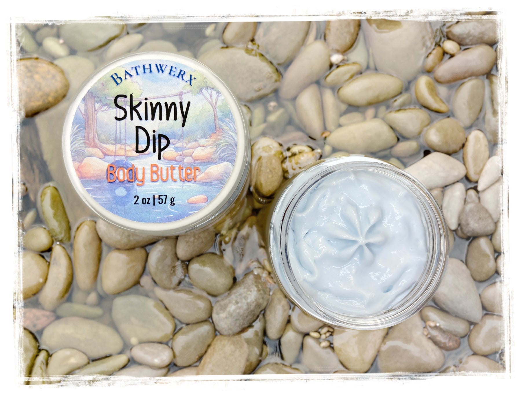 BathWerx Skinny Dip Body Butter open clear jar showing light blue creamy texture, on smooth river stones with water