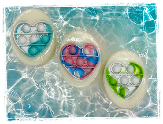 Three handcrafted kids' soaps with embedded silicone pop-it fidget toys in teal, pink, and green, displayed on a rippling water background