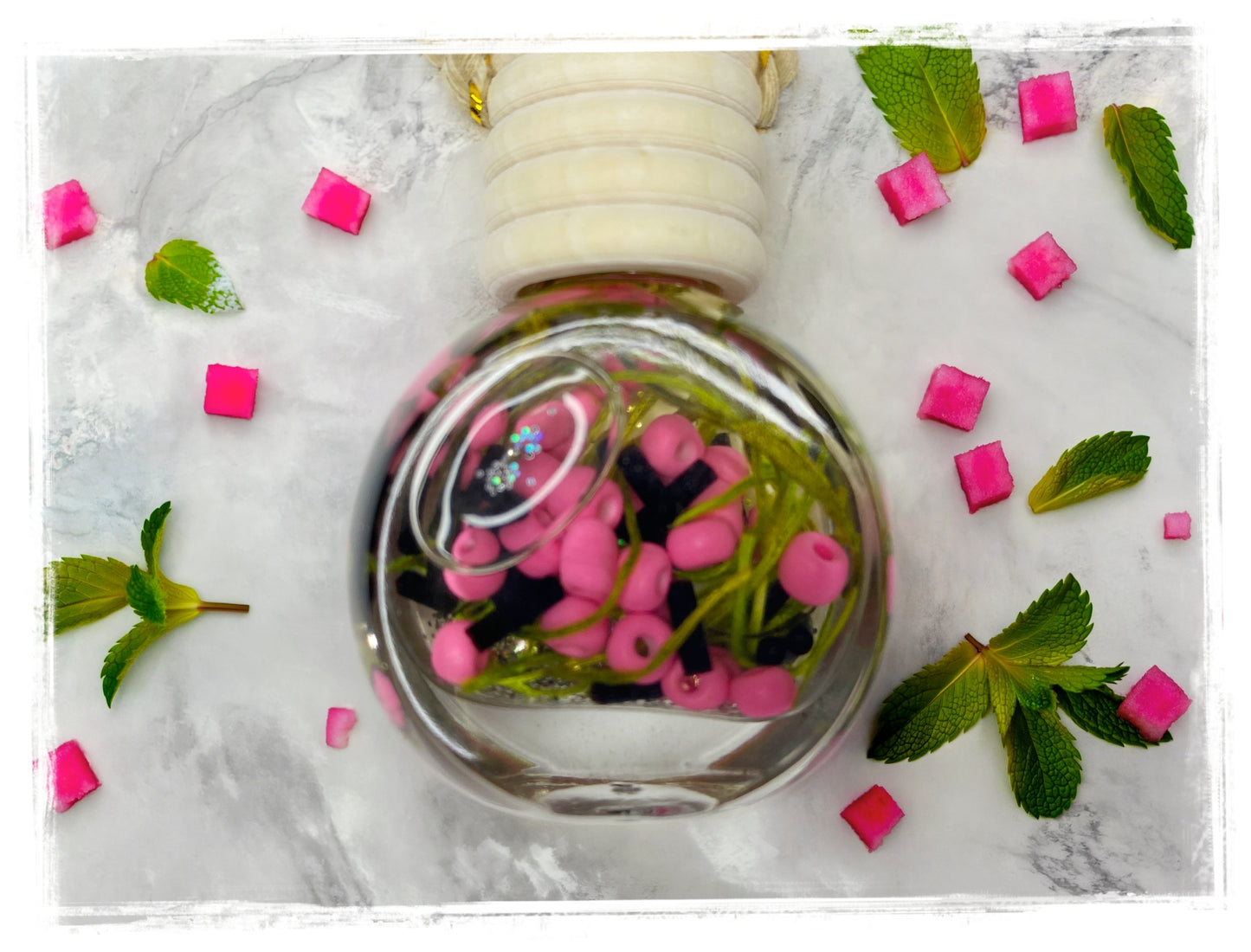 BathWerx Watermelon Mint Car Charm — glass globe pendant filled with pink beads, black accents, green botanicals, and glitter, with a white cap and gold hook, styled on marble with fresh mint leaves and pink candy pieces