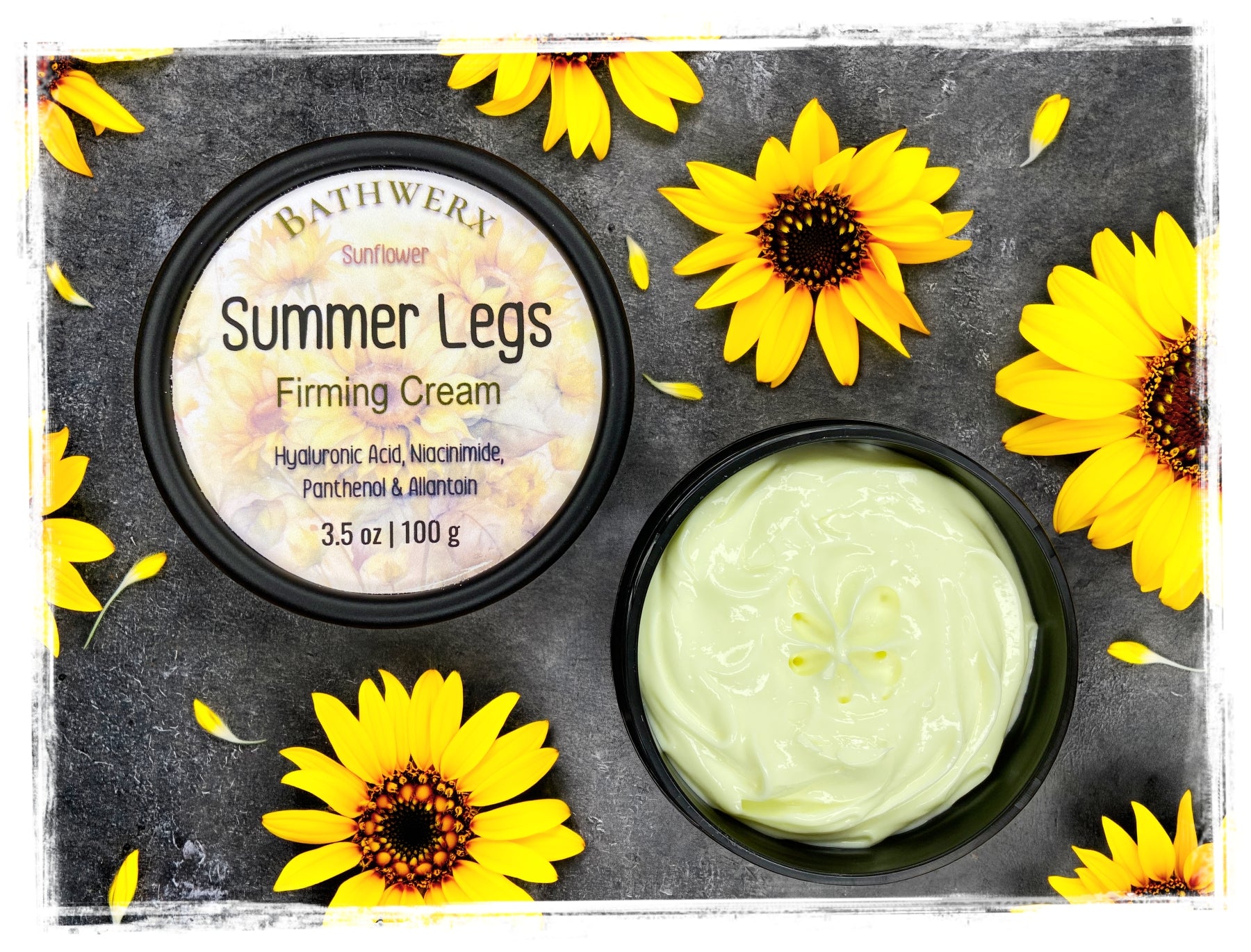 BathWerx Summer Legs Firming Cream open jar and lid with sunflower label, surrounded by fresh yellow sunflowers on dark slate — hyaluronic acid, niacinamide, panthenol, 3.5 oz.