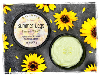 BathWerx Summer Legs Firming Cream open jar and lid with sunflower label, surrounded by fresh yellow sunflowers on dark slate — hyaluronic acid, niacinamide, panthenol, 3.5 oz.