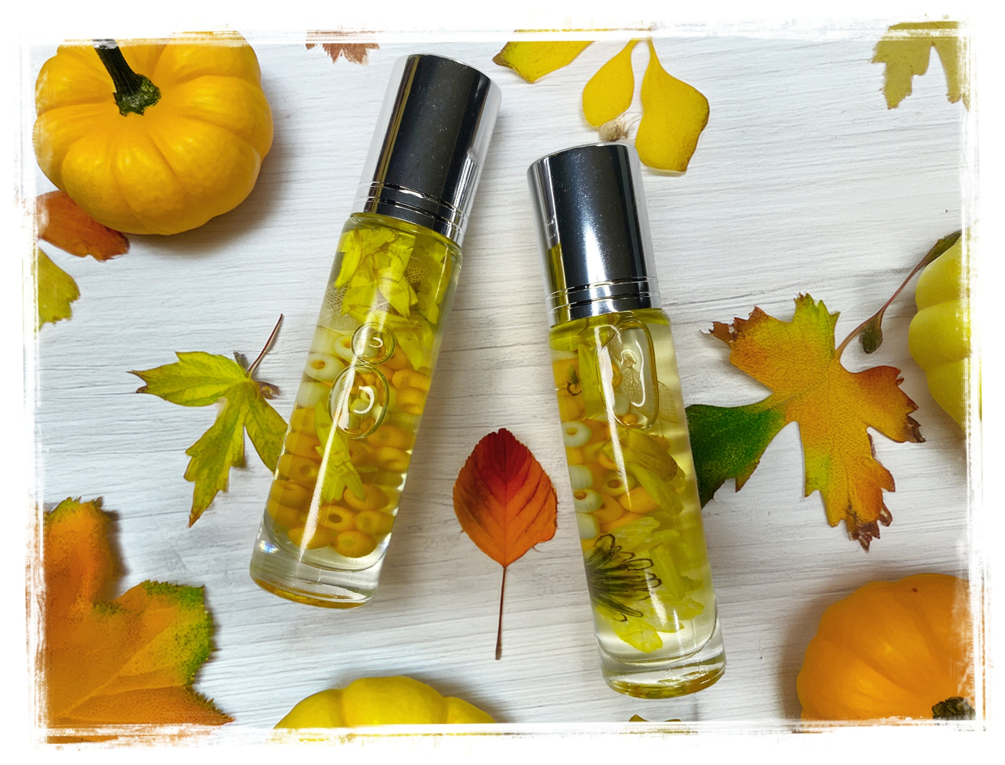 Two BathWerx Autumn Magic Roll-on Perfume bottles with silver caps, filled with golden oil, yellow botanicals, and beads, styled on a white wood surface with mini pumpkins and colorful fall leaves