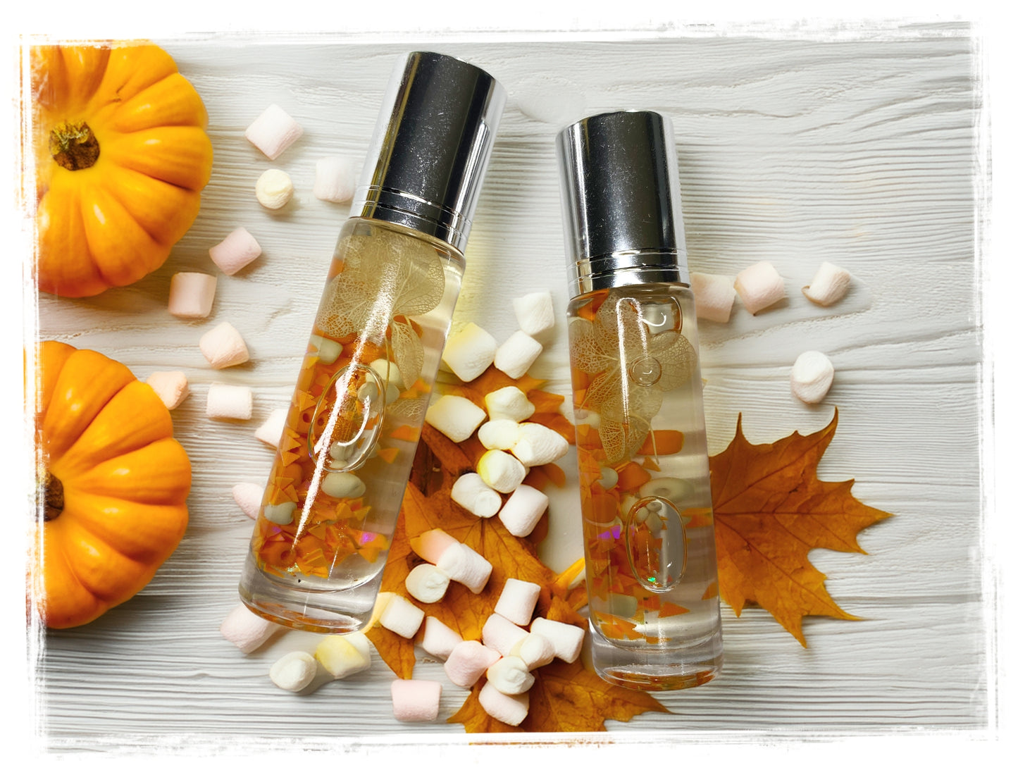 Two BathWerx Vanilla Marshmallow Pumpkin Roll-on Perfume bottles with silver caps, filled with golden oil and orange botanicals, resting on dried maple leaves surrounded by mini marshmallows and orange pumpkins on a white wood surface