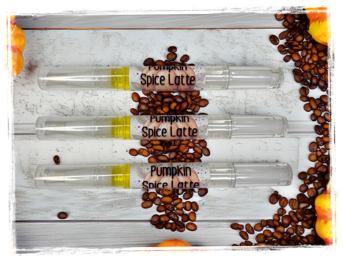 Three Pumpkin Spice Latte Moisturizing Nail Pens displayed horizontally on a white wood surface, surrounded by coffee beans and pumpkins