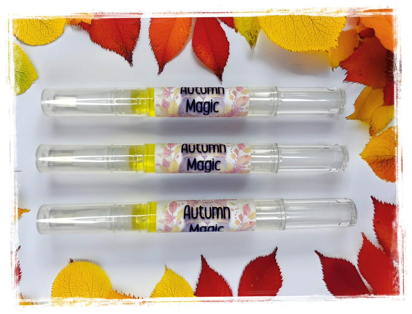 Three Autumn Magic Moisturizing Nail Pens displayed horizontally on a white surface, surrounded by vibrant red, orange, and yellow autumn leaves
