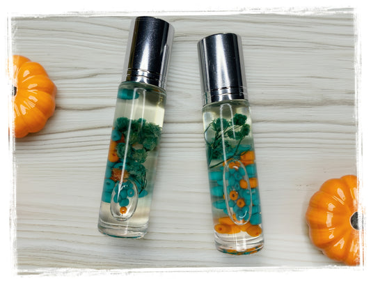 Two Forbidden Pumpkin Fall Roll-On Perfume Oil bottles with silver caps on a white wood surface, filled with clear oil containing teal flowers and orange beads, flanked by orange mini pumpkins