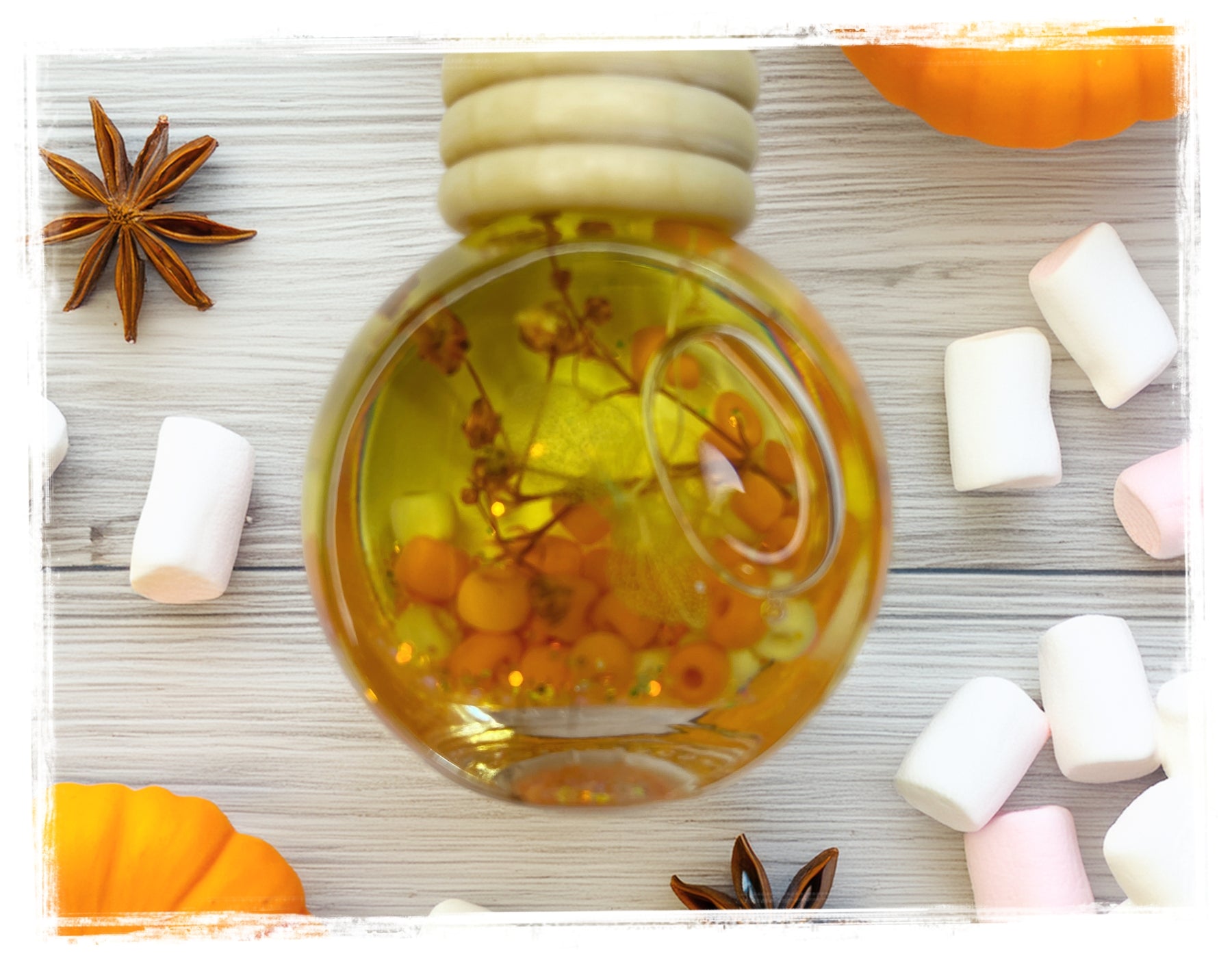 Marshmallow Pumpkin Fall Scented Car Charm - close-up of a round glass charm filled with golden oil, orange beads, and dried botanicals, surrounded by mini marshmallows, star anise, and mini pumpkins on a white wood surface.