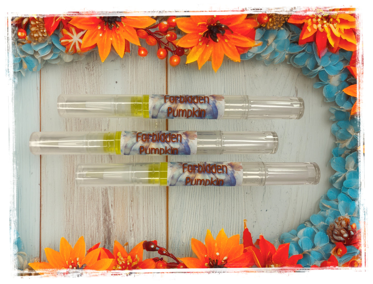 Three Forbidden Pumpkin Nail & Cuticle Repair Pens arranged on a rustic white wood surface, surrounded by vibrant orange and blue fall flowers with pinecones and red berries