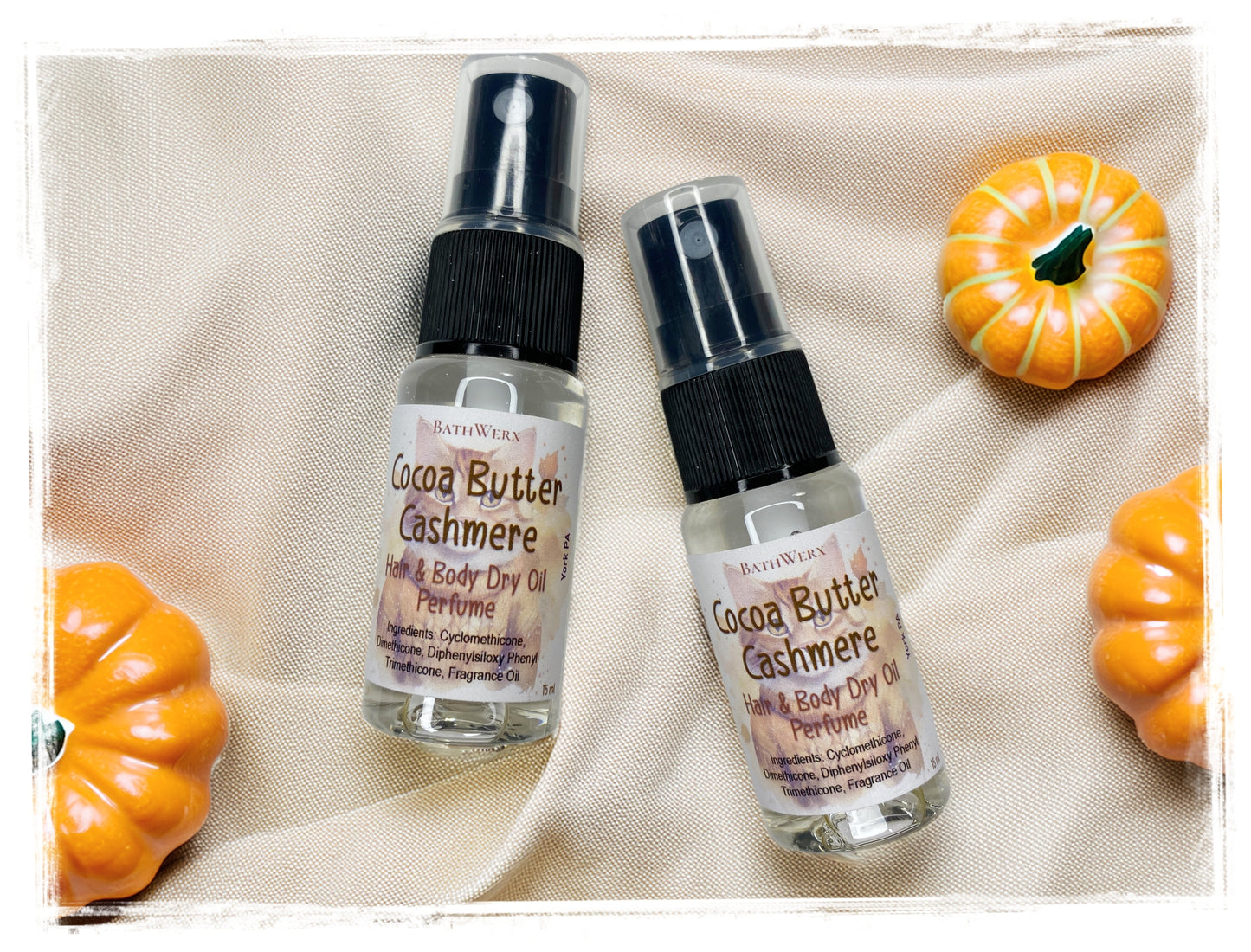 Two BathWerx Cocoa Butter Cashmere Hair & Body Dry Oil Perfume spray bottles with clear glass and black pump tops, styled on a linen cloth with orange decorative pumpkins