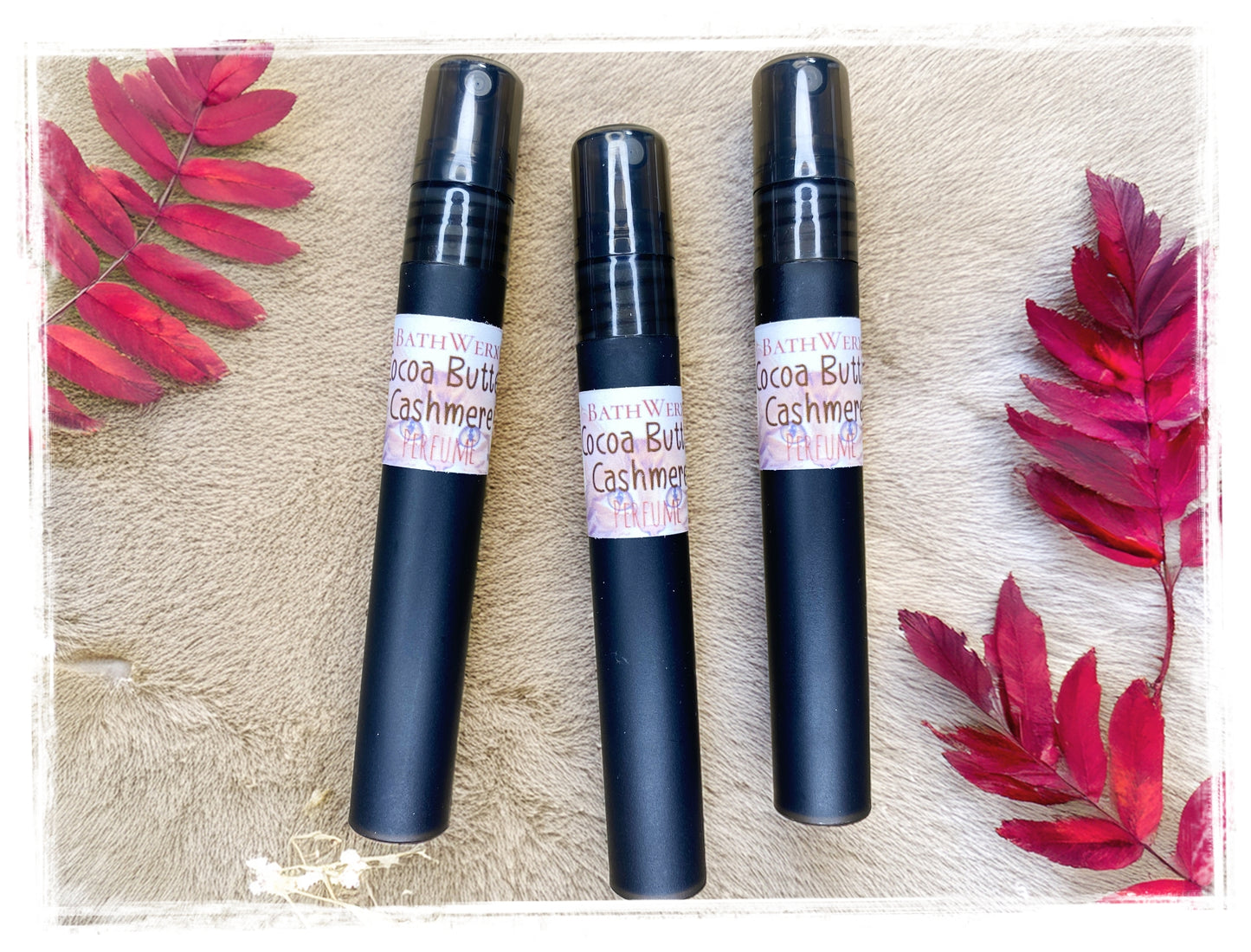 BathWerx Cocoa Butter Cashmere perfume sprays in matte black, styled on a soft cream fur surface with vibrant red autumn leaves - travel-size 10ml fall fragrance
