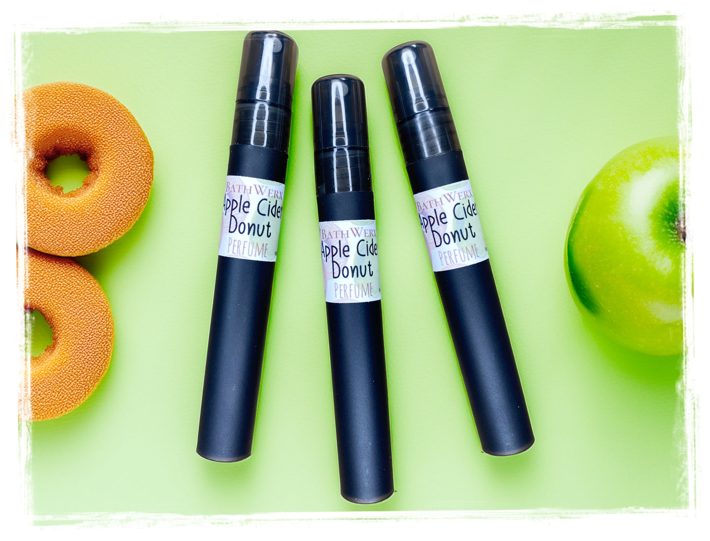 Three BathWerx Apple Cider Donut perfume mist spray vials in matte black, arranged on a light green background with apple cider donuts and a green apple.