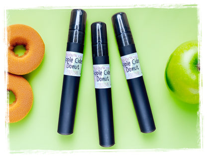 Three BathWerx Apple Cider Donut perfume mist spray vials in matte black, arranged on a light green background with apple cider donuts and a green apple.