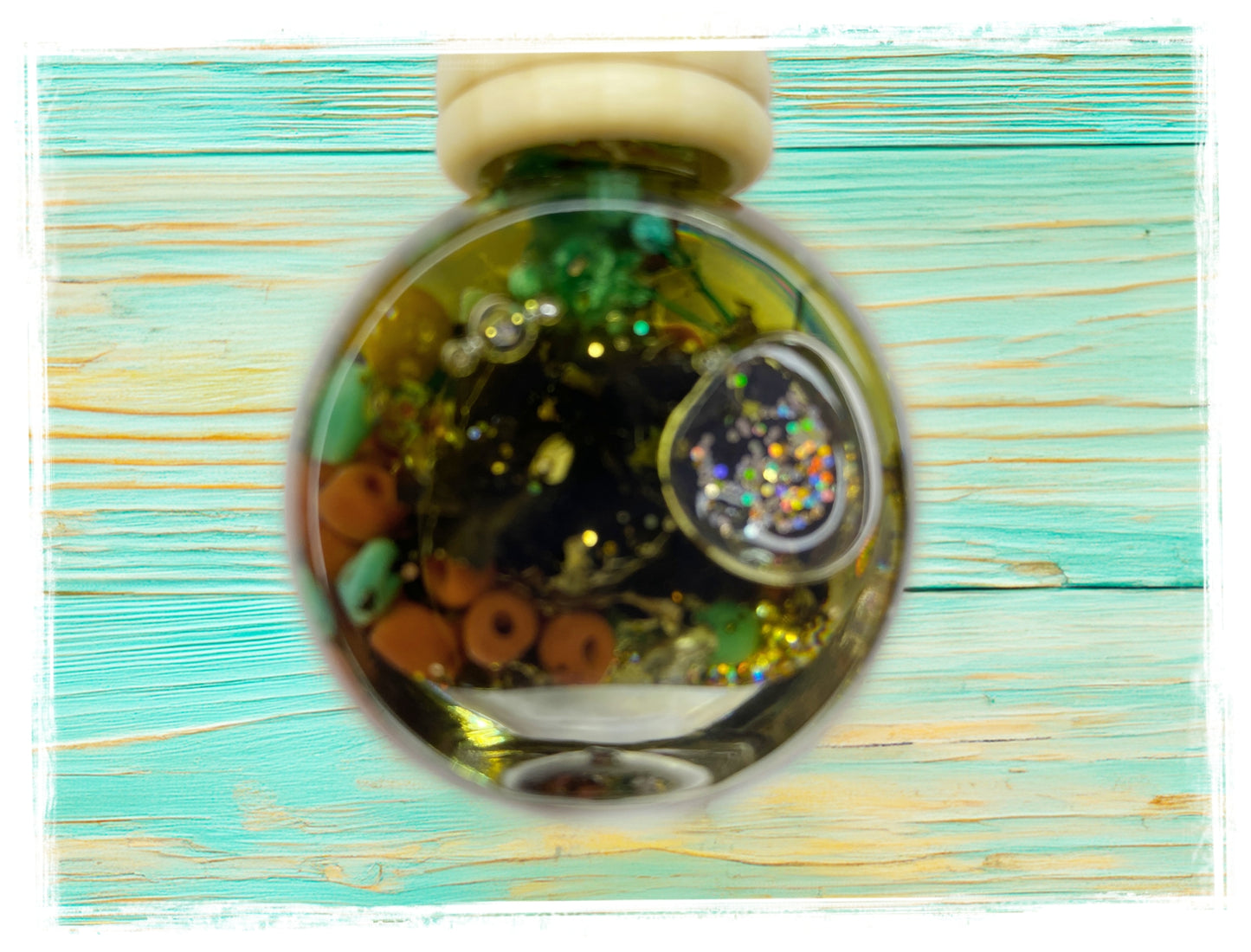 Close-up of a BathWerx Maple Bacon car charm globe filled with gold glitter, green botanicals, orange beads, and a holographic gem, hanging against a teal wood background