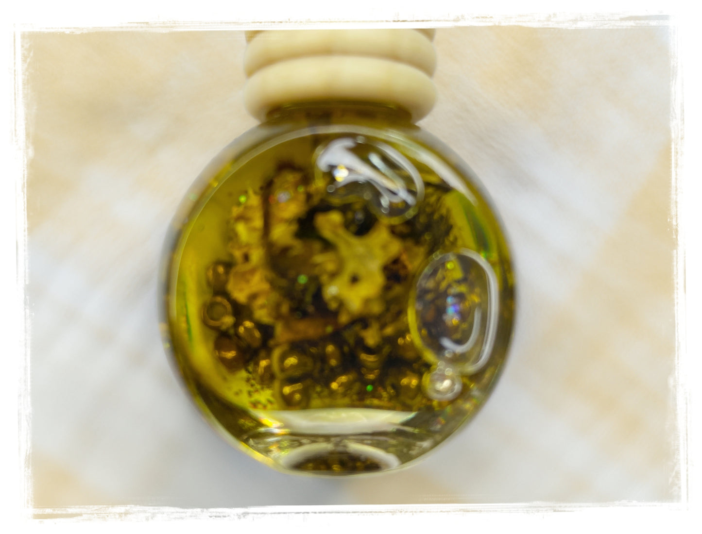 Close-up of a BathWerx Glazed Pecans car charm globe filled with golden oil, amber botanicals, and a holographic gem, hanging against a soft cream background