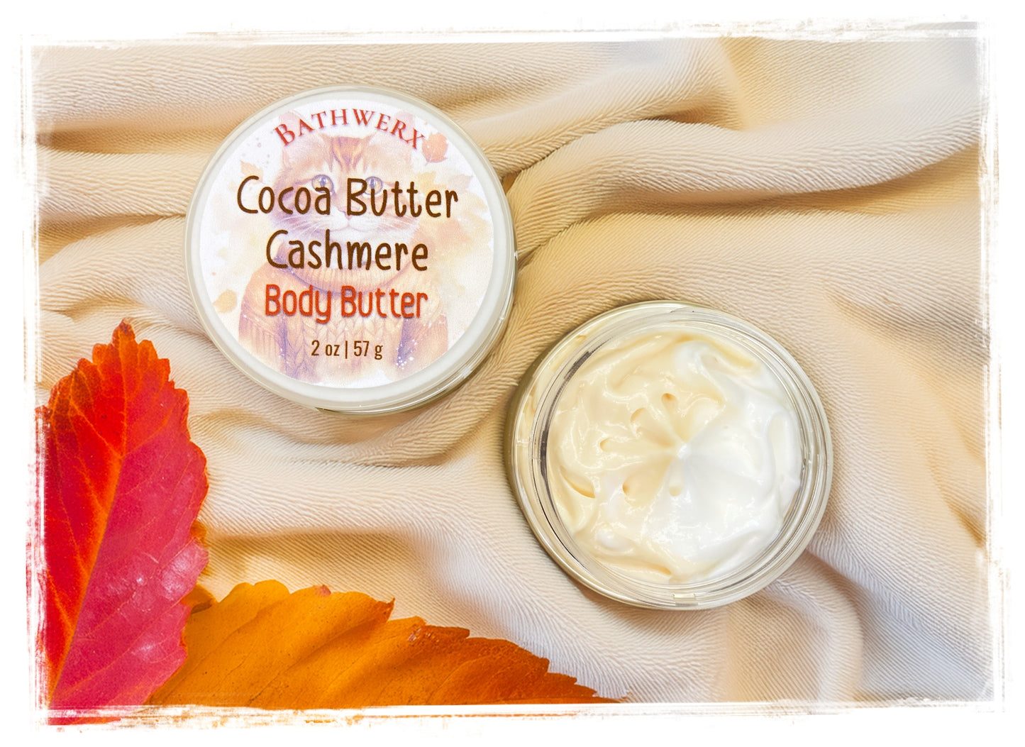 BathWerx Cocoa Butter Cashmere Body Butter jar open to reveal creamy white whipped texture, styled on a soft cashmere fabric with vibrant red and orange autumn leaves - 2 oz / 57g