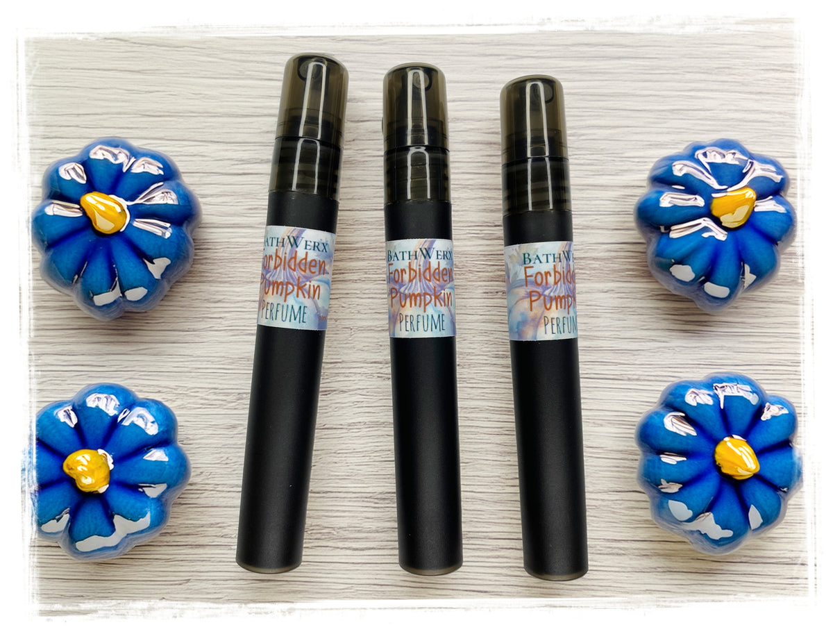 Three BathWerx Forbidden Pumpkin Perfume spray pens in black, displayed on a light wood surface surrounded by blue ceramic flower decorations.