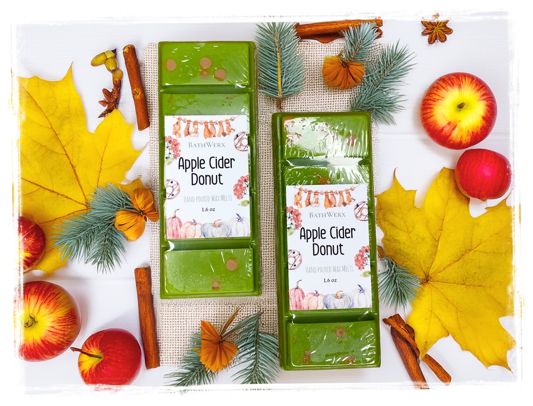 Two BathWerx Apple Cider Donut hand-poured wax melts in sage green, displayed on a white surface with burlap, surrounded by red apples, yellow maple leaves, pine sprigs, cinnamon sticks, star anise, and dried orange flowers.