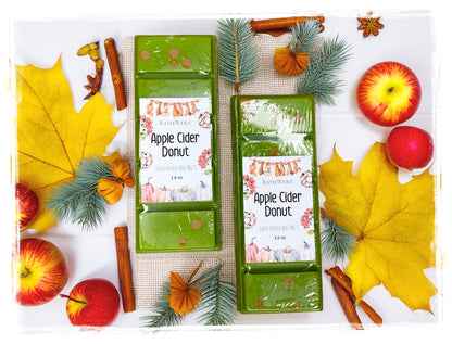 Two BathWerx Apple Cider Donut hand-poured wax melts in sage green, displayed on a white surface with burlap, surrounded by red apples, yellow maple leaves, pine sprigs, cinnamon sticks, star anise, and dried orange flowers.