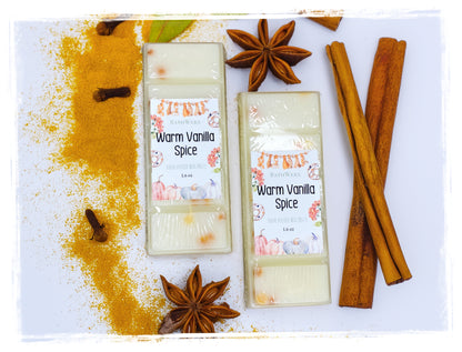 Two BathWerx Warm Vanilla Spice hand-poured wax melts in creamy white, displayed on a white surface with a dramatic sweep of golden turmeric spice powder, cinnamon sticks, star anise, and whole cloves.