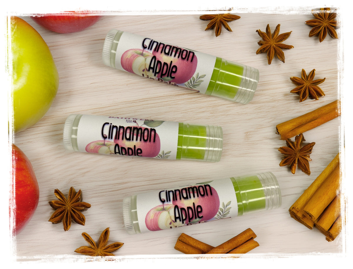 BathWerx Cinnamon Apple Lip Balm tubes on a white wood surface with red and green apples, cinnamon sticks, and star anise.