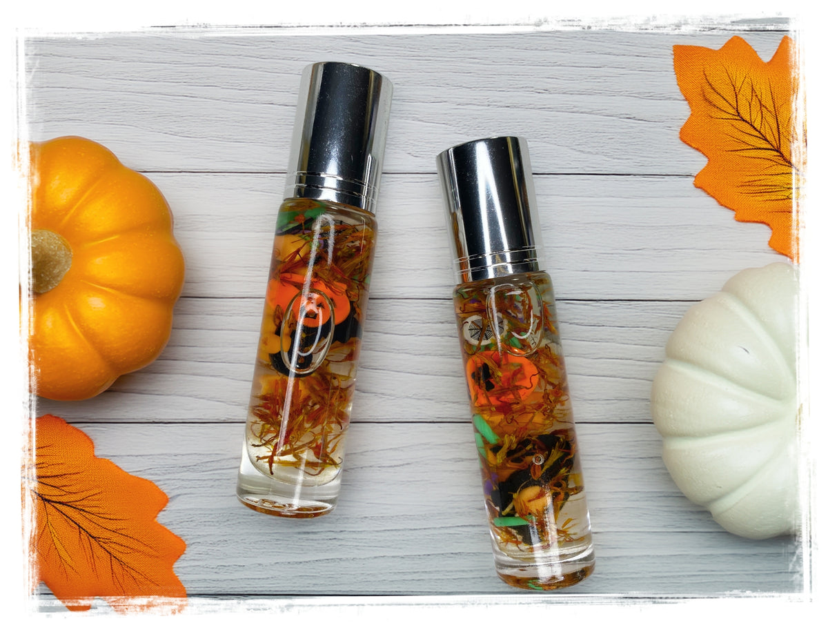 Two BathWerx Toasted Pumpkin roll-on perfume oil bottles filled with amber oil and vibrant autumn botanicals, displayed on a white wood surface alongside an orange pumpkin, white pumpkin, and fall leaves.