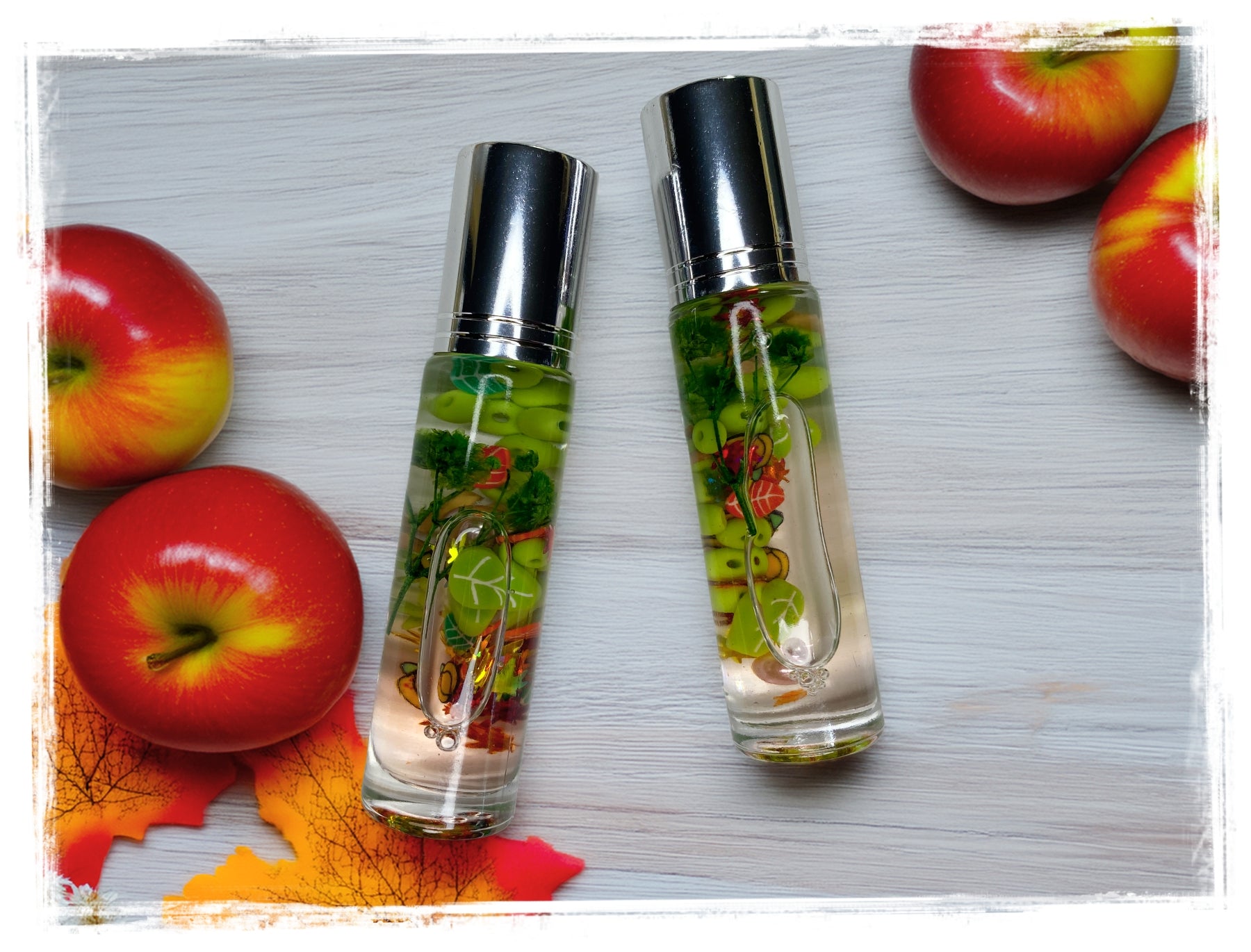 Two clear roll-on perfume oil bottles filled with light oil and vibrant green botanicals, displayed on a white wood surface surrounded by red apples and colorful fall leaves.
