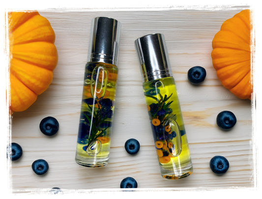 Two BathWerx Blueberry Pumpkin roll-on perfume oil bottles filled with golden oil, deep blue botanicals, and orange beads, displayed on a white wood surface flanked by orange pumpkins and scattered fresh blueberries.