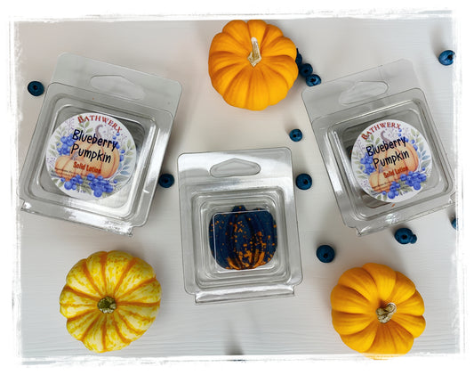 BathWerx Blueberry Pumpkin solid lotion bar in clear clamshell packaging, center bar shown unwrapped revealing a deep navy blue lotion bar with gold speckles, displayed with mini pumpkins and fresh blueberries on a white wood surface.