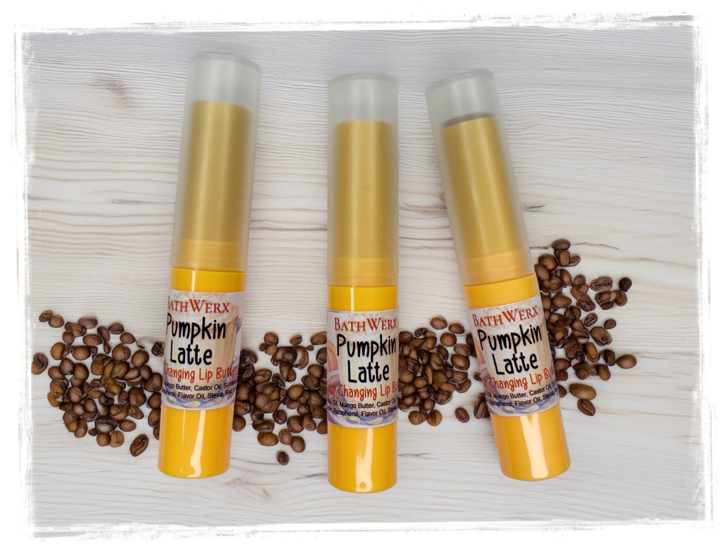 Three BathWerx Pumpkin Latte Color-Changing Lip Butter tubes in gold packaging surrounded by scattered coffee beans on a white wood background.