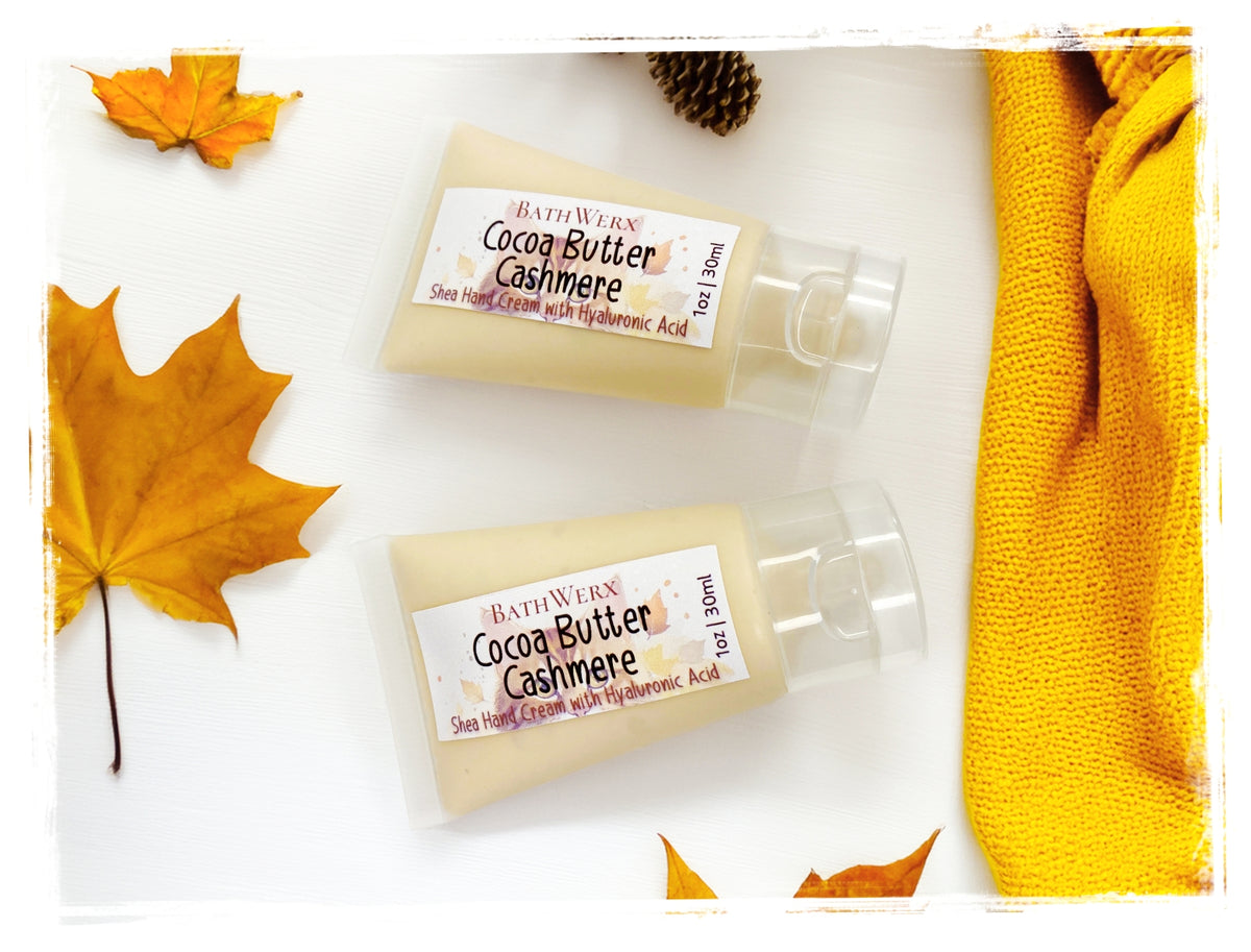 BathWerx Cocoa Butter Cashmere Shea Hand Cream with Hyaluronic Acid, 1oz travel-size pump bottles styled with autumn leaves and a golden knit scarf.