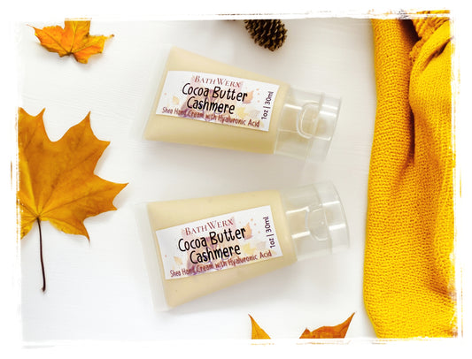BathWerx Cocoa Butter Cashmere Shea Hand Cream with Hyaluronic Acid, 1oz travel-size pump bottles styled with autumn leaves and a golden knit scarf.