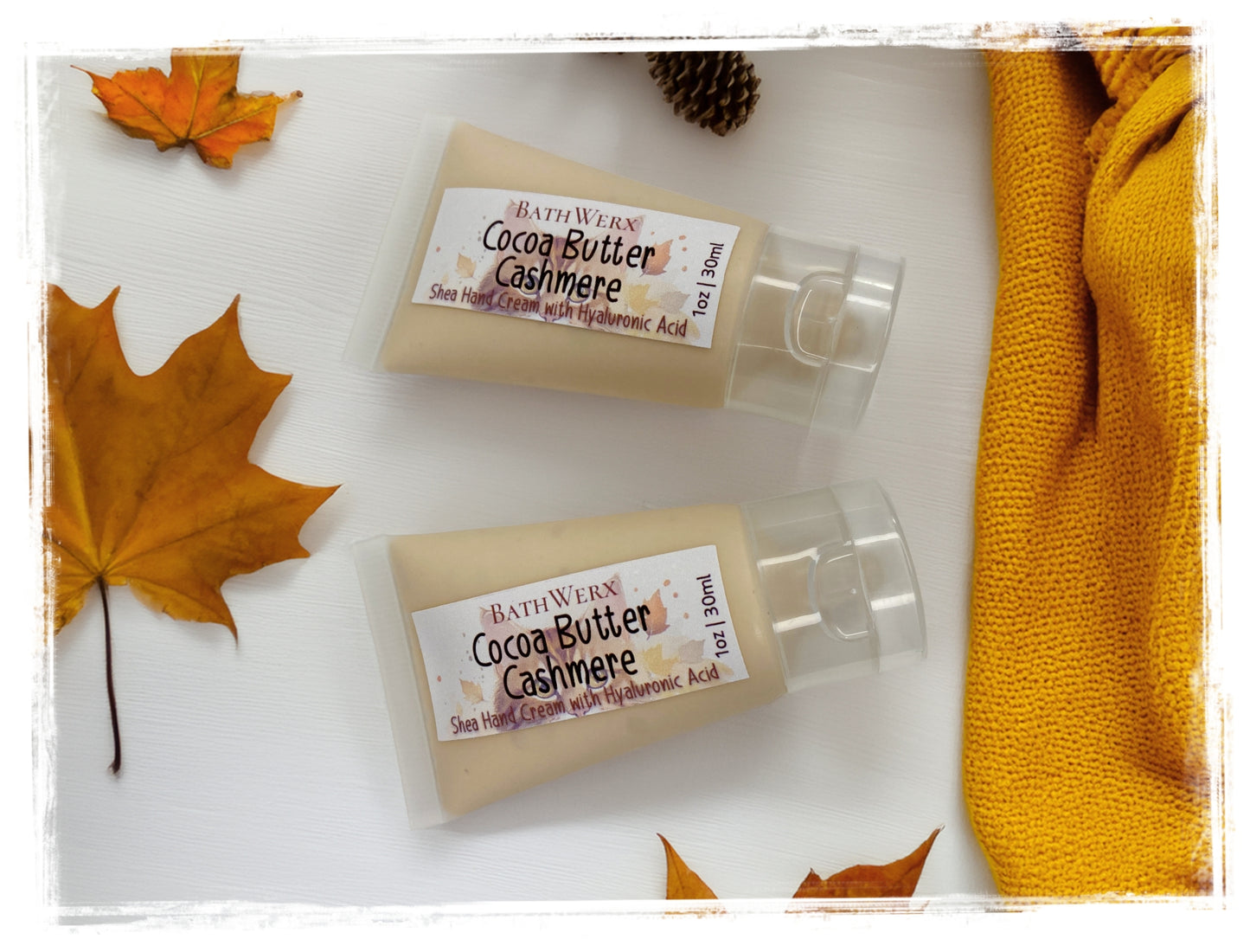BathWerx Cocoa Butter Cashmere Shea Hand Cream with Hyaluronic Acid, 1oz travel-size pump bottles styled with autumn leaves and a golden knit scarf.