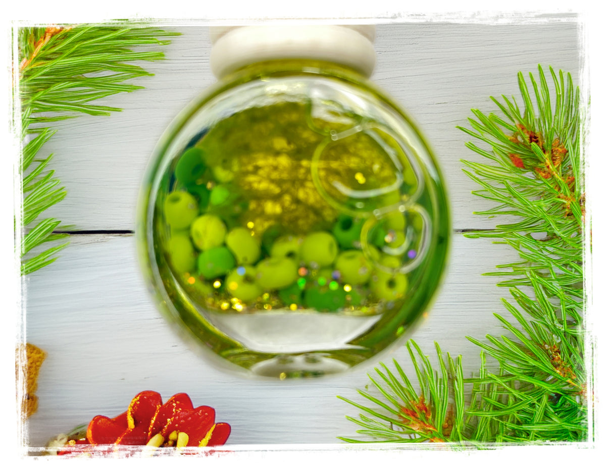 BathWerx Christmas Tree Car Charm — clear glass ornament filled with green beads, gold glitter, and coiled wire accents, displayed on a white wood surface with fresh pine branches and a red holiday decoration.