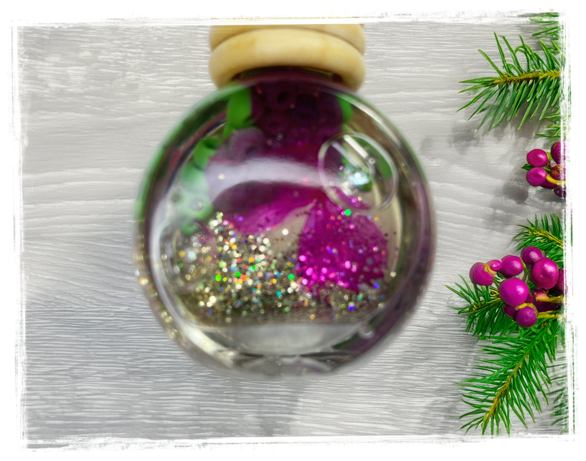 BathWerx Bayberry Birch Car Charm — clear glass ornament filled with magenta rose petals, silver and holographic glitter, and a bubble accent, displayed on a white wood surface with fresh pine branches and pink holiday berries.