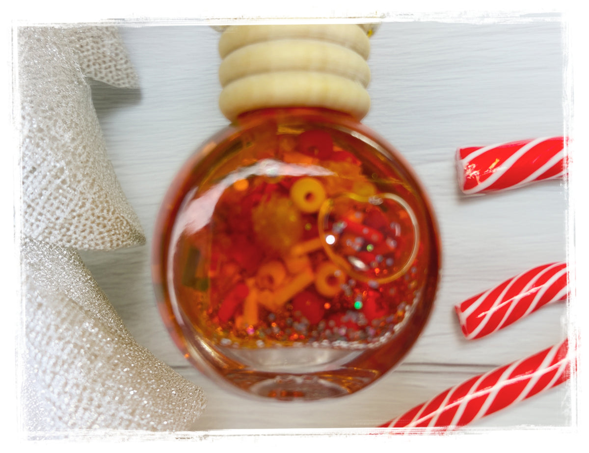 BathWerx Peppermint Milkshake Car Charm — clear glass ornament filled with red and orange confetti, coiled wire accents, and multicolored glitter, displayed on a white wood surface with candy cane pieces and a silver ribbon.