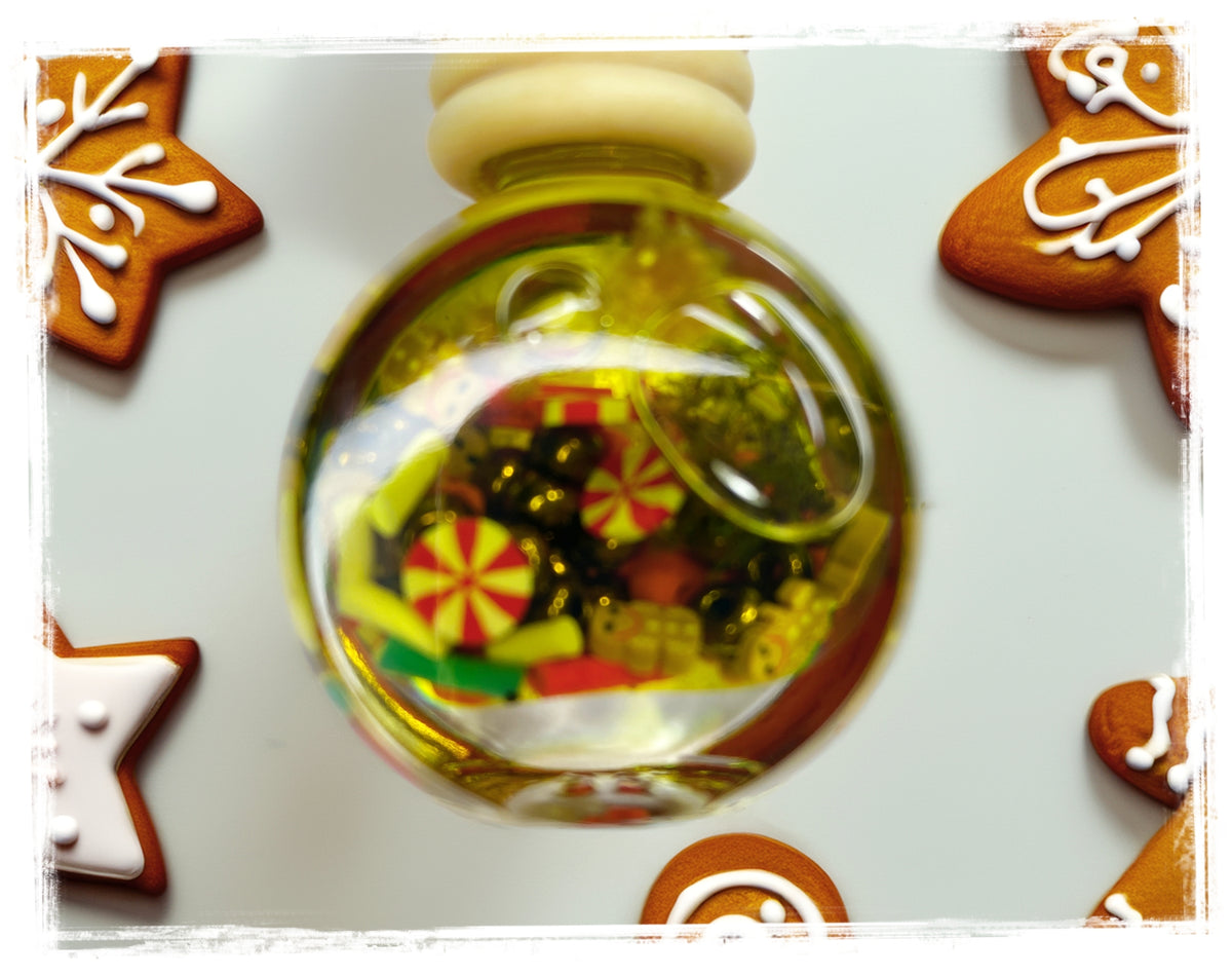 BathWerx Gingerbread Car Charm — clear glass ornament filled with yellow-green oil, miniature peppermint candy charms, dark beads, colorful confetti, and a coiled wire accent, displayed on a light blue surface surrounded by iced gingerbread star cookies.
